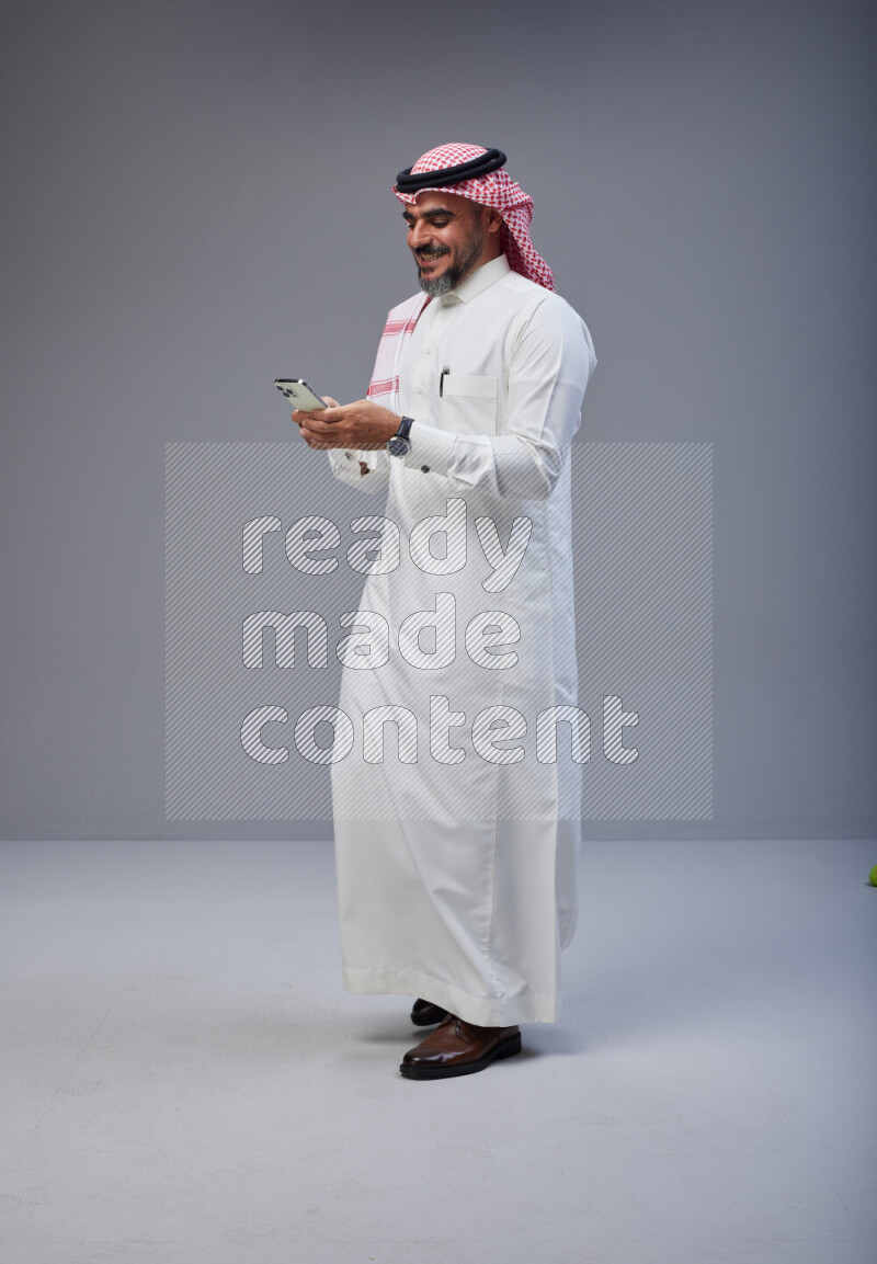 Saudi man Wearing Thob and red Shomag standing texting on phone on Gray background