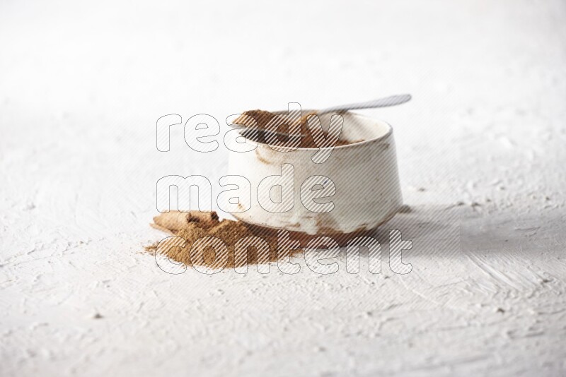 Ceramic beige bowl full of cinnamon powder and a metal spoon with cinnamon sticks next of it on a textured white background