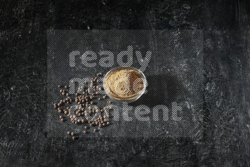 وعاء زجاجي ممتلئ ببودرة بهار الفلفل الحلو وحبوبه متناثرة على خلفية سوداء