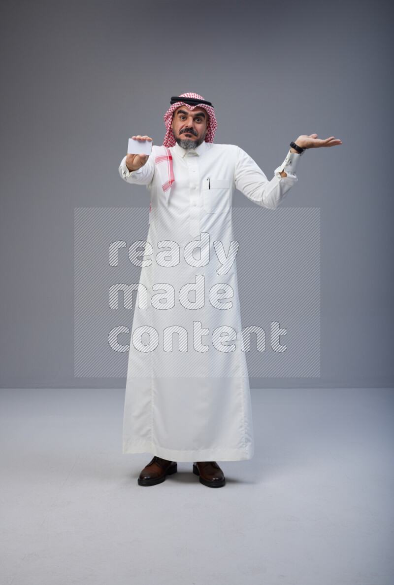 Saudi man Wearing Thob and red Shomag standing holding ATM card on Gray background