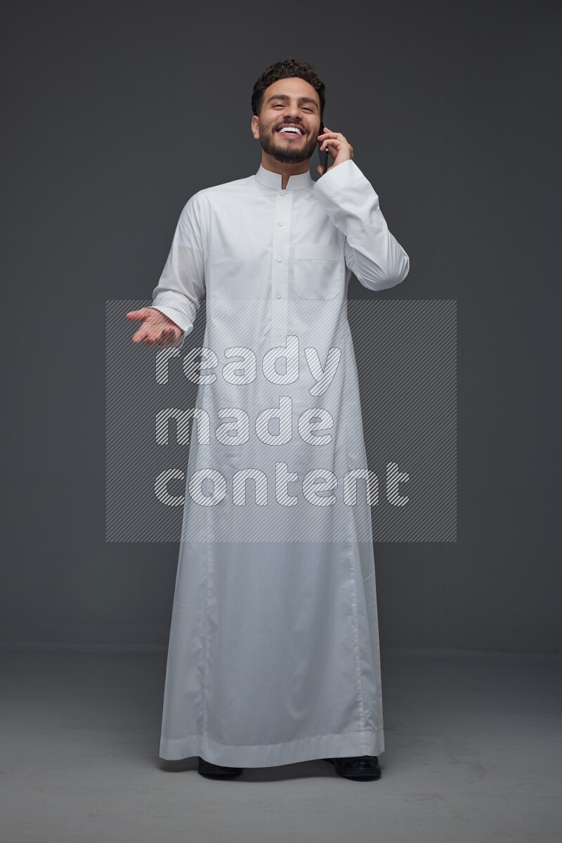 A Saudi man wearing Thobe and talking in the phone while standing and making different poses eye level on a gray background