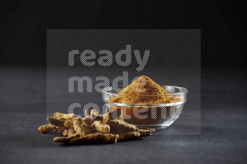 A glass bowl full of turmeric powder and dried whole fingers beside it on black flooring