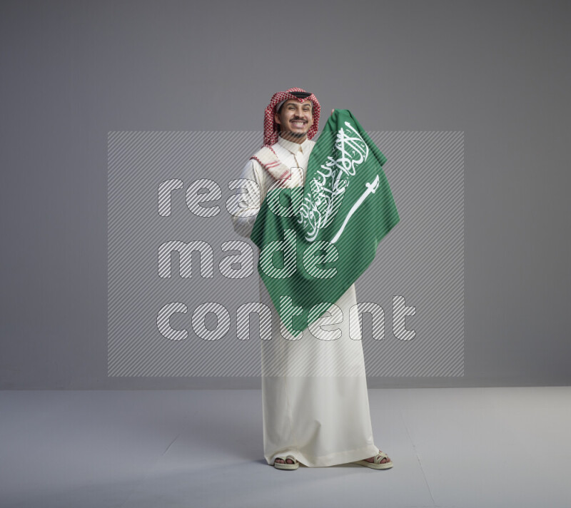 A saudi man standing wearing thob and red shomag holding big saudi flag on gray background