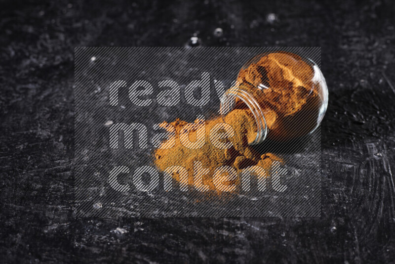A glass jar full of ground paprika powder flipped with some spilling powder on black background