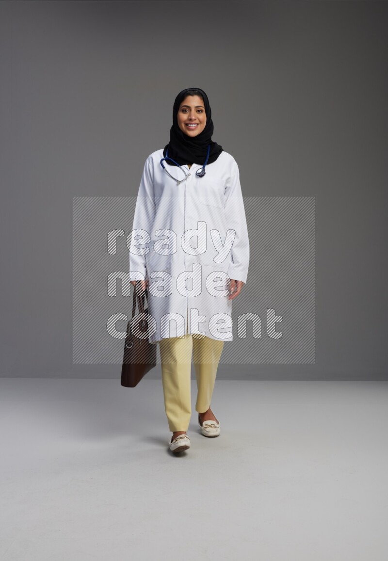 Saudi woman wearing lab coat with stethoscope standing holding bag on Gray background