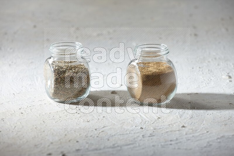 2 glass spice jars full of cumin powder and cumin seeds on textured white flooring