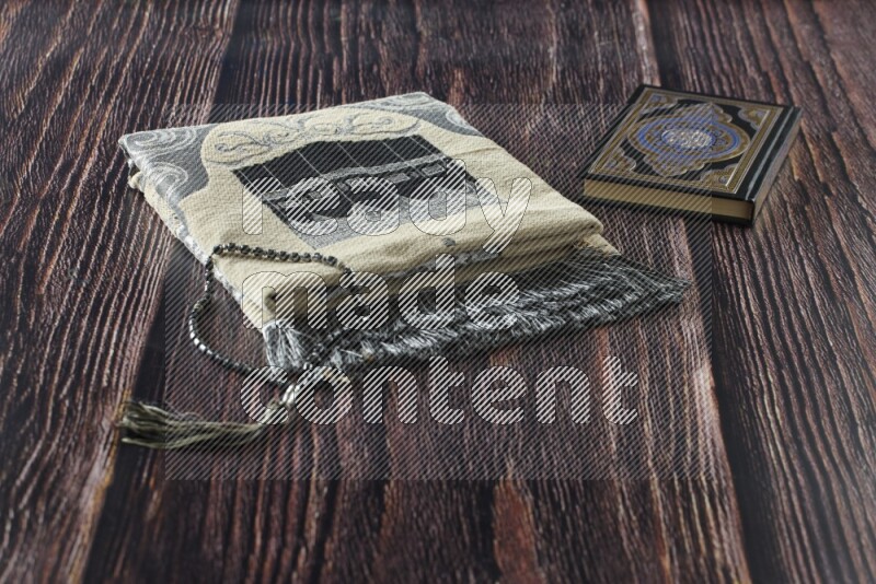 A folded prayer mat with different elements such as quran and prayer beads on brown wooden background