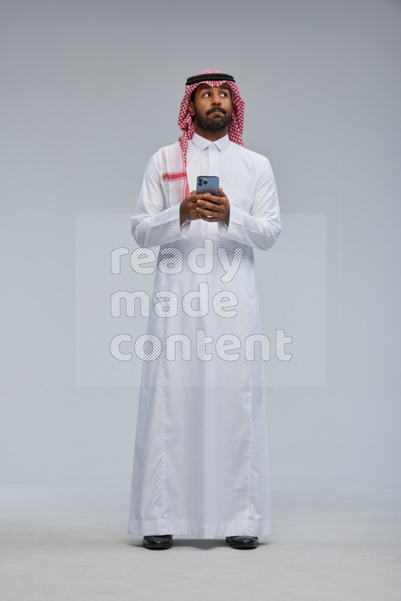 Saudi man Wearing Thob and shomag standing texting on phone on Gray background