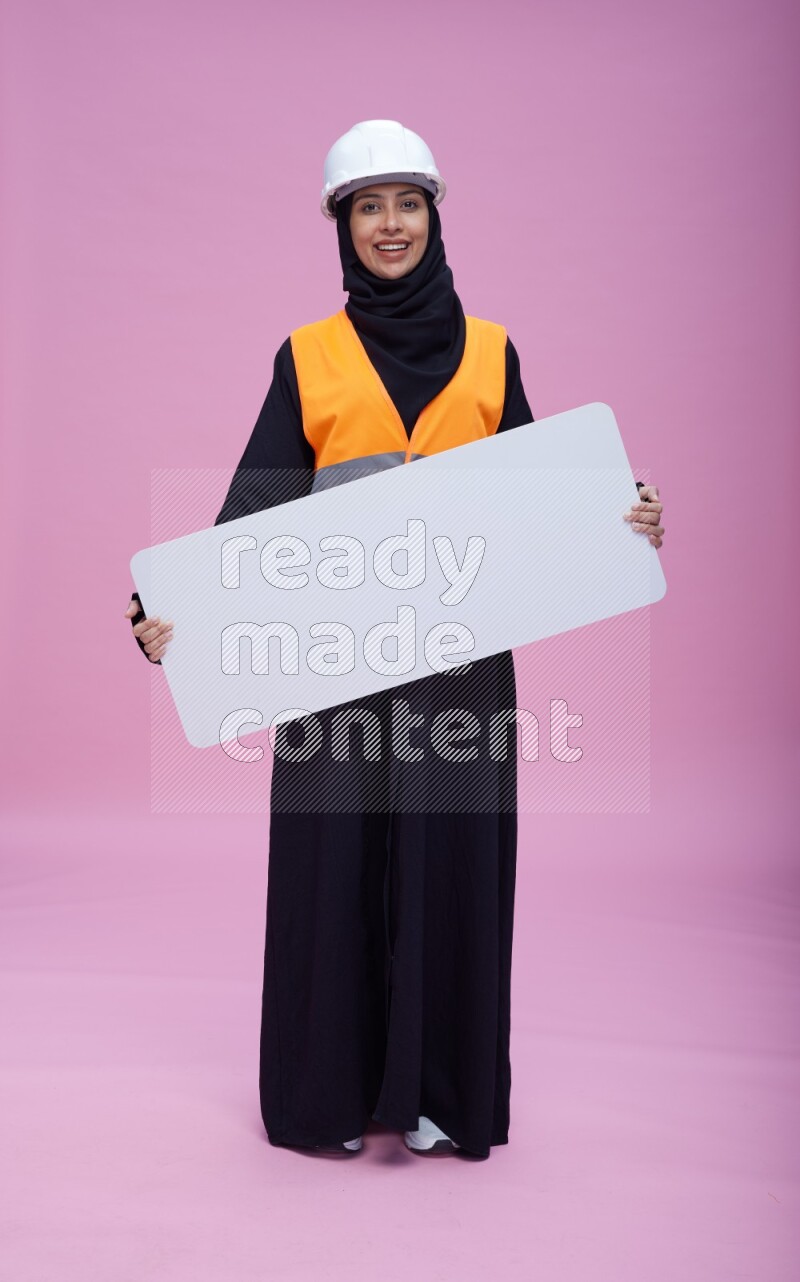 Saudi woman wearing Abaya with engineer vest and helmet standing holding board on pink background