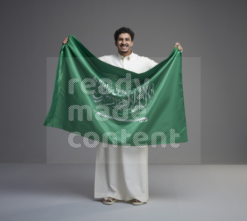 A Saudi man standing wearing thob holding big Saudi flag on gray background