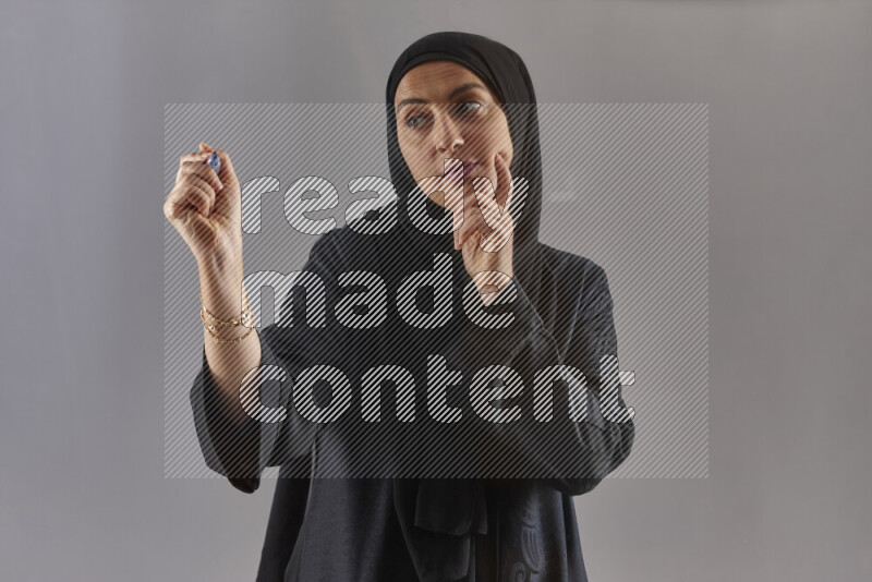 Woman in black abaya and hijab holding a marker pen to write on a transparent board with different reactions and expressions on grey background