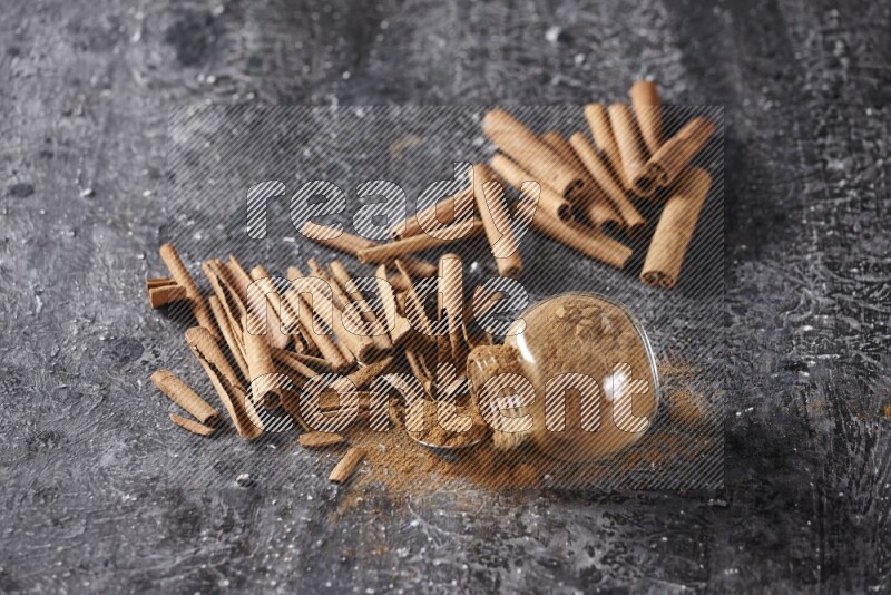 Herbal glass jar full cinnamon powder flipped and a metal spoon full of powder surrounded by cinnamon sticks on textured black background in different angles