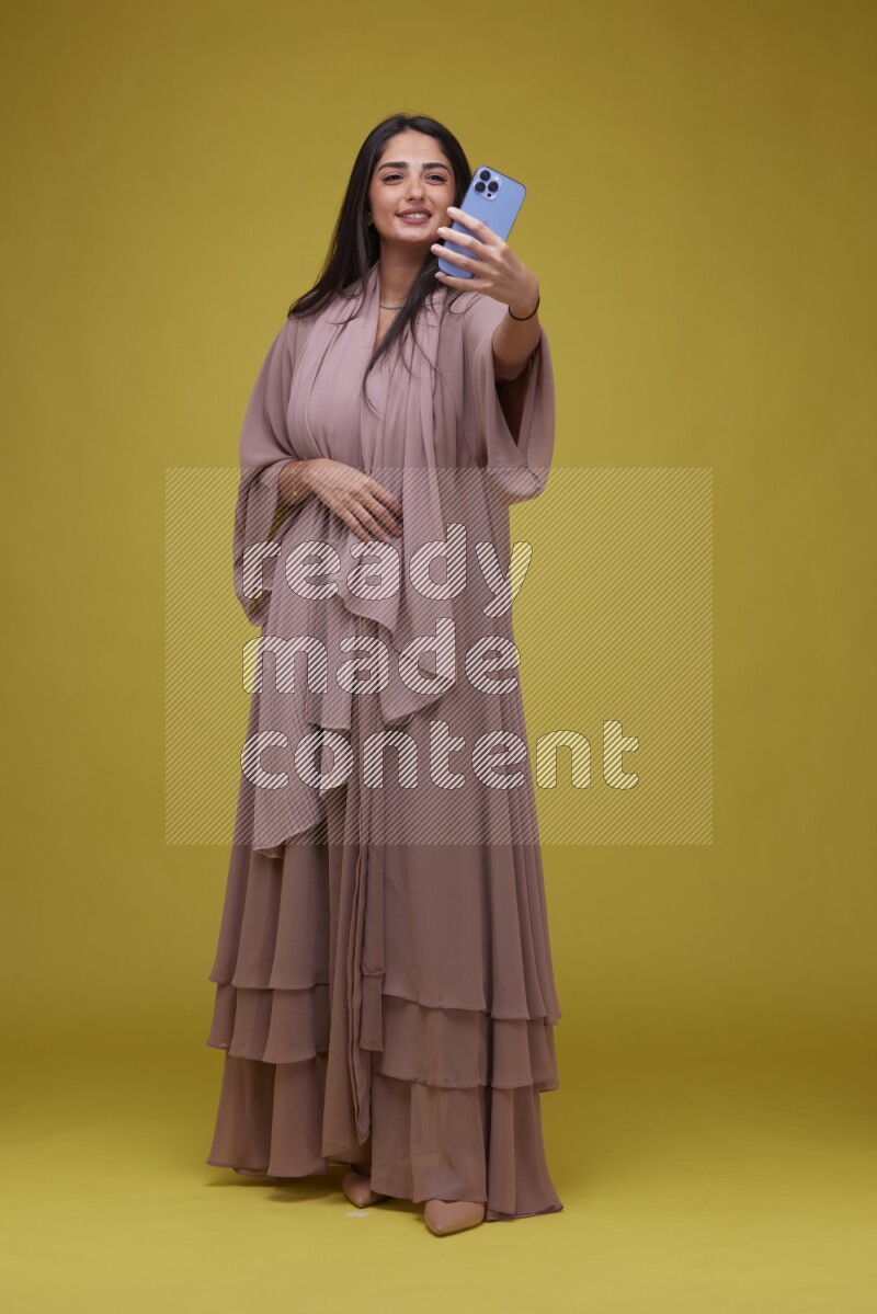 A woman Taking a Selfie on a Yellow Background wearing Brown Abaya