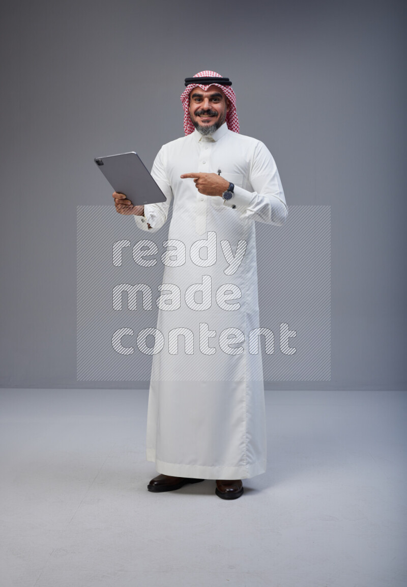 Saudi man Wearing Thob and red Shomag standing working on tablet on Gray background