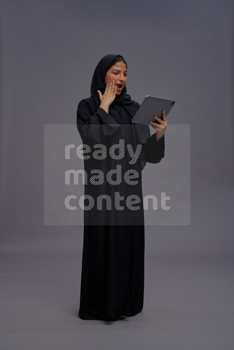 Saudi woman wearing Abaya standing working on tablet on gray background
