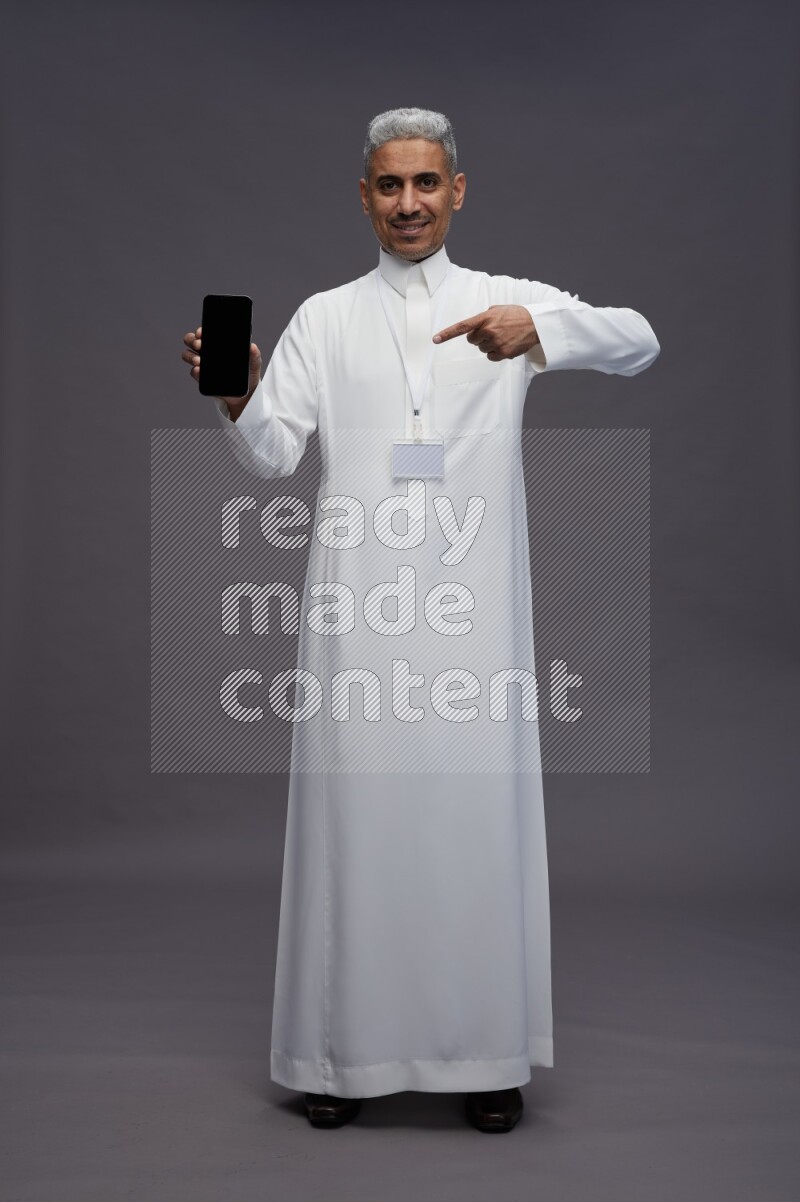 Saudi man wearing thob with neck strap employee badge standing showing phone to camera on gray background