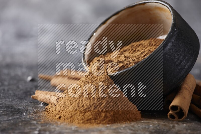 Black pottery bowl over filled with cinnamon powder and cinnamon sticks around the bowl on a textured black background