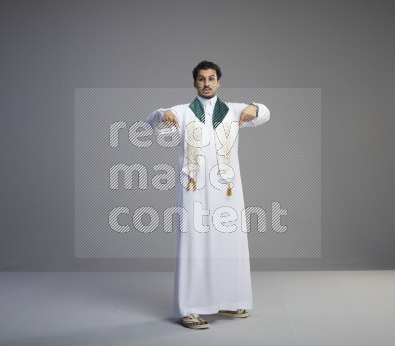 A Saudi man standing wearing thob and Saudi flag scarf with face painting on gray background