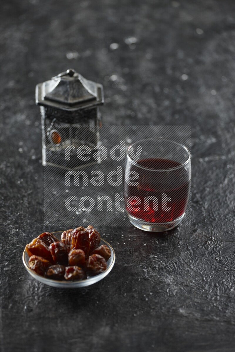 A silver lantern with different drinks, dates, nuts, prayer beads and quran on textured black background