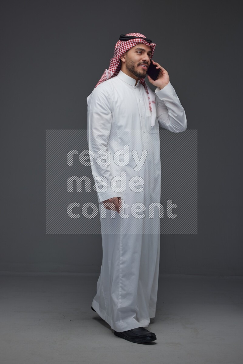 A Saudi man wearing Thobe and Shmagh talking in the phone eye level on a gray background