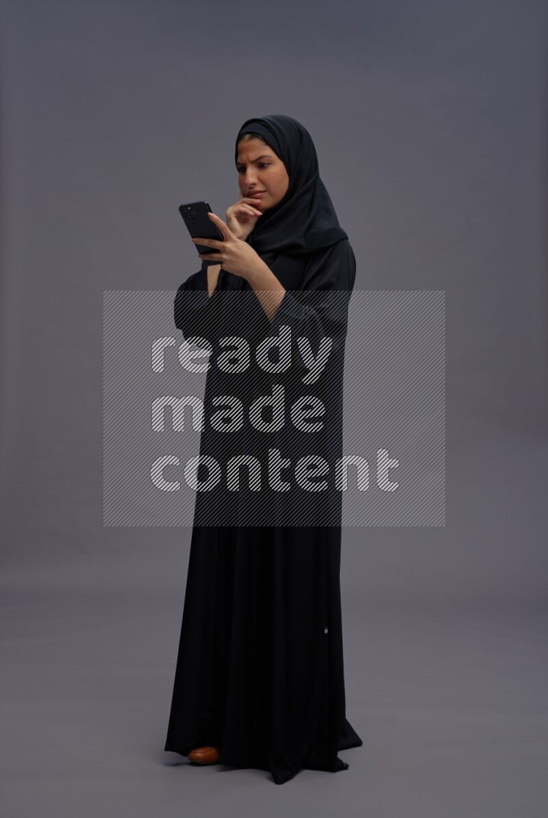 Saudi woman wearing Abaya standing texting on phone on gray background