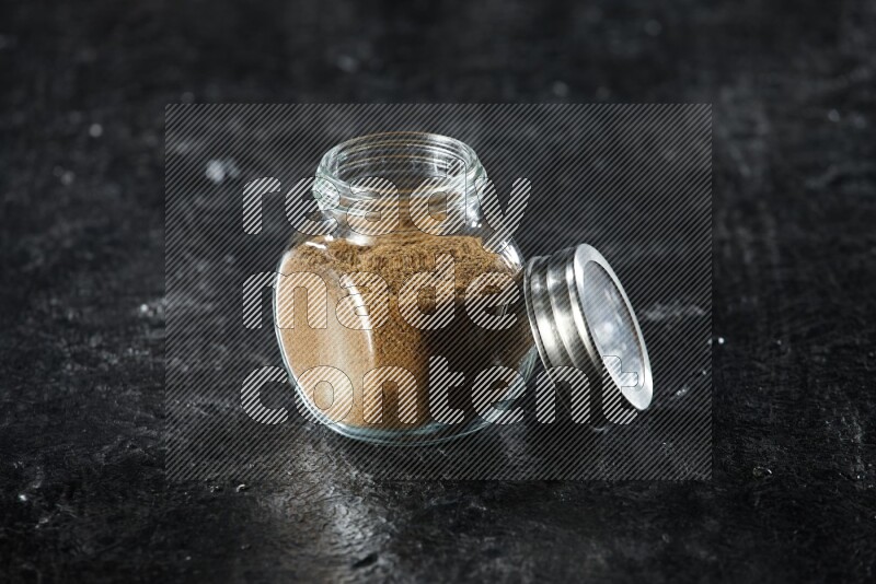 A glass spice jar full of cumin powder on a textured black flooring