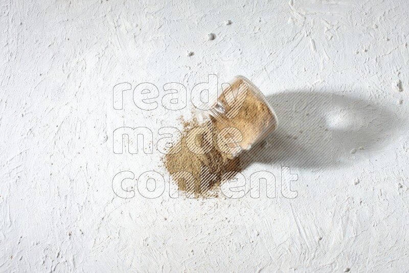 وعاء زجاجي مقلوب ممتلئ ببودرة الكمون والبودرة تناثرت منه على خلفية بيضاء