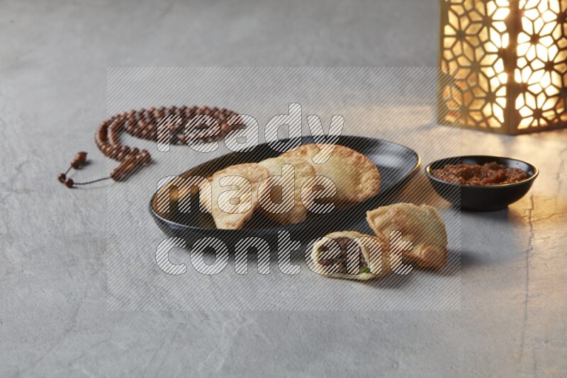 Three fried sambosas in an oval shaped black plate and a red sauce in a black round ramekin with a brown misbaha and a golden lantern on the side on a gray background