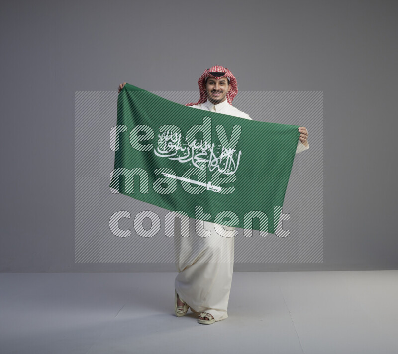 A saudi man standing wearing thob and red shomag holding big saudi flag on gray background