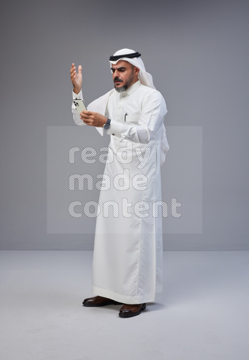 Saudi man Wearing Thob and white Shomag standing texting on phone on Gray background
