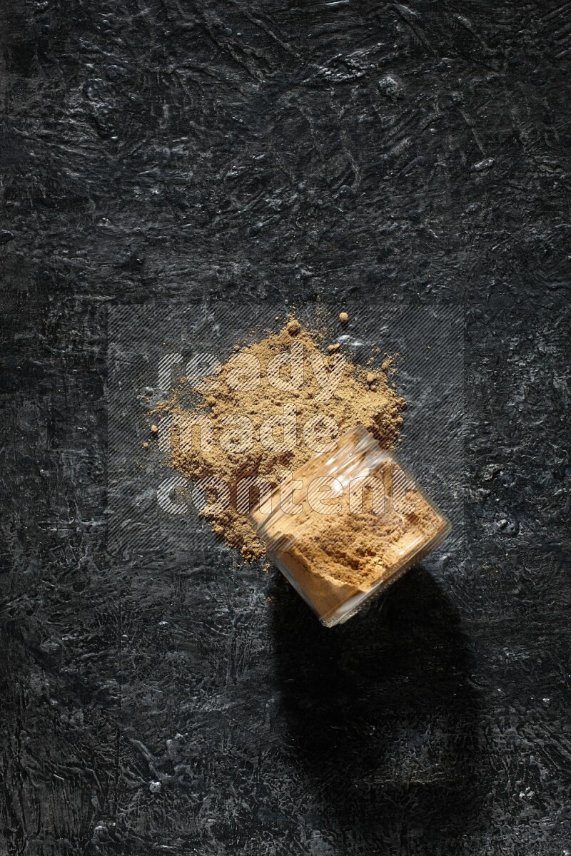 A flipped glass jar full of allspice powder and powder spilled out of it on a textured black flooring