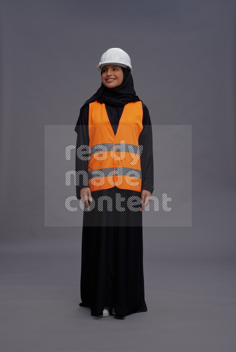 Saudi woman wearing Abaya with engineer vest standing interacting with the camera on gray background