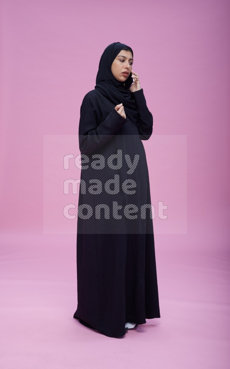 Saudi woman wearing Abaya standing talking on phone on pink background