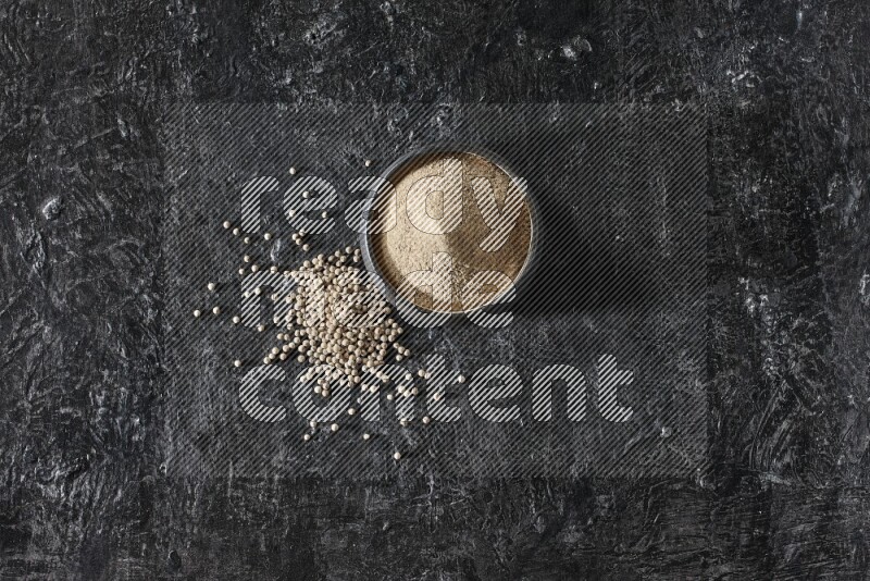 Black pottery bowl full of white pepper powder with white pepper beads on textured black flooring