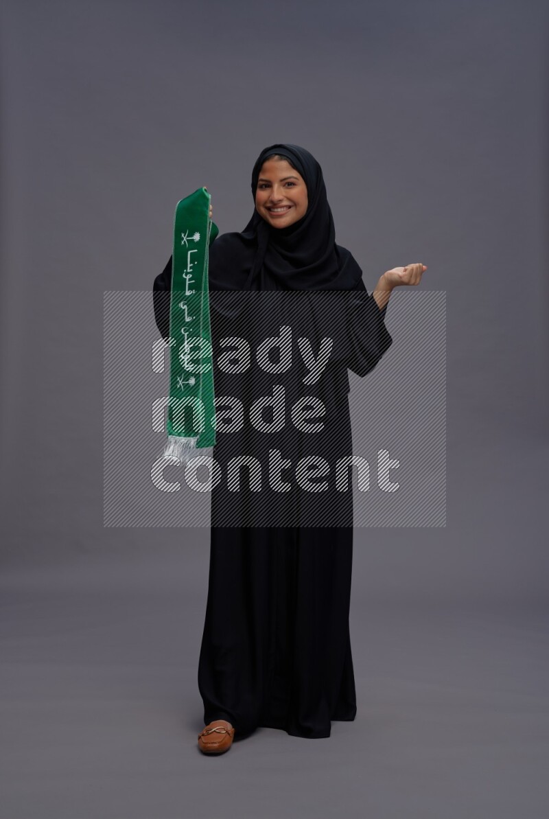 Saudi woman wearing Abaya standing holding Saudi flag on gray background