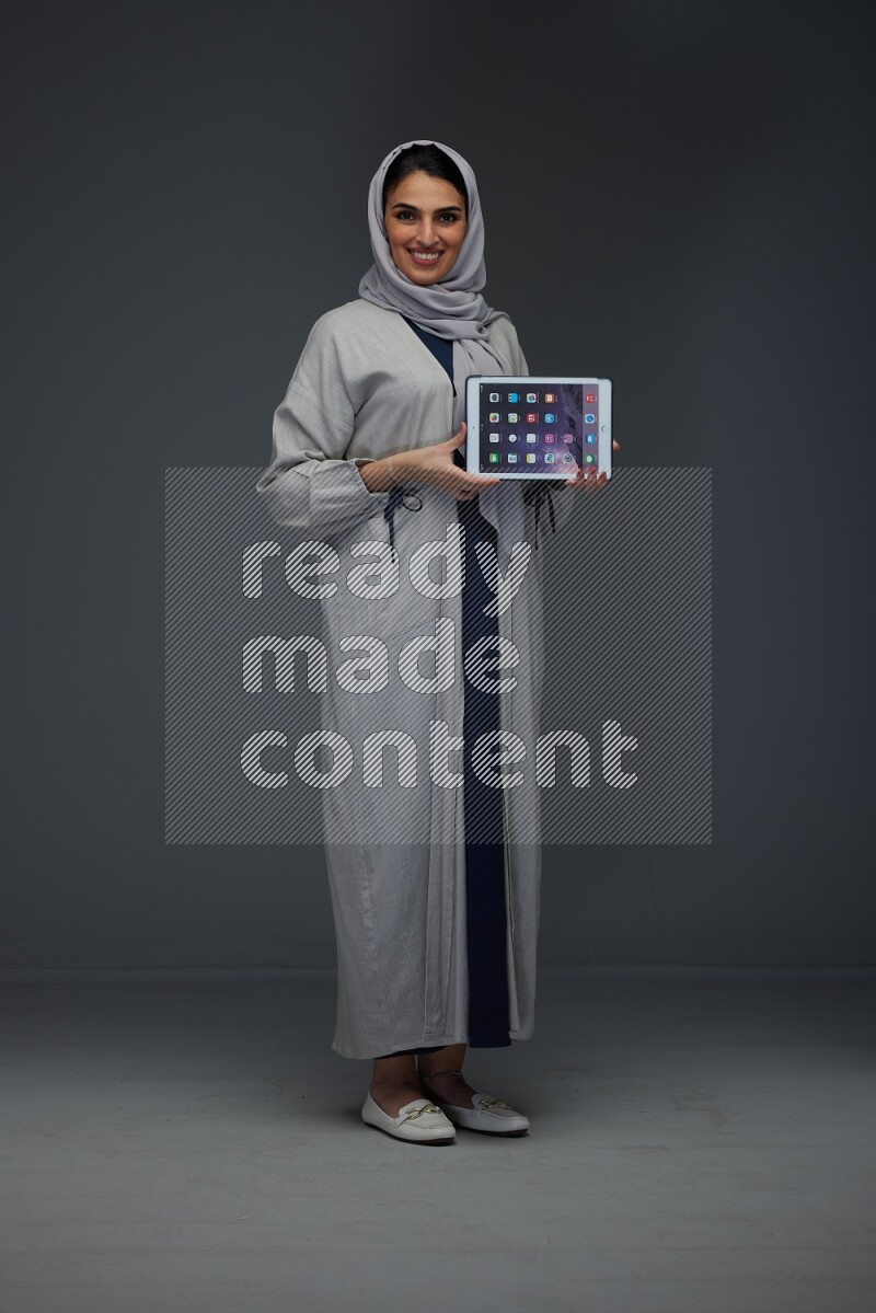A Saudi woman wearing a light gray Abaya and head scarf standing and showing the phone's screen eye level on a grey background