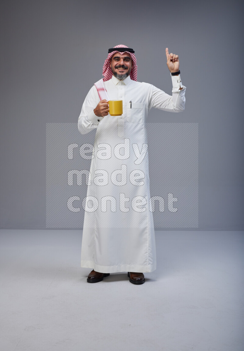 Saudi man Wearing Thob and red Shomag standing holding mug on Gray background