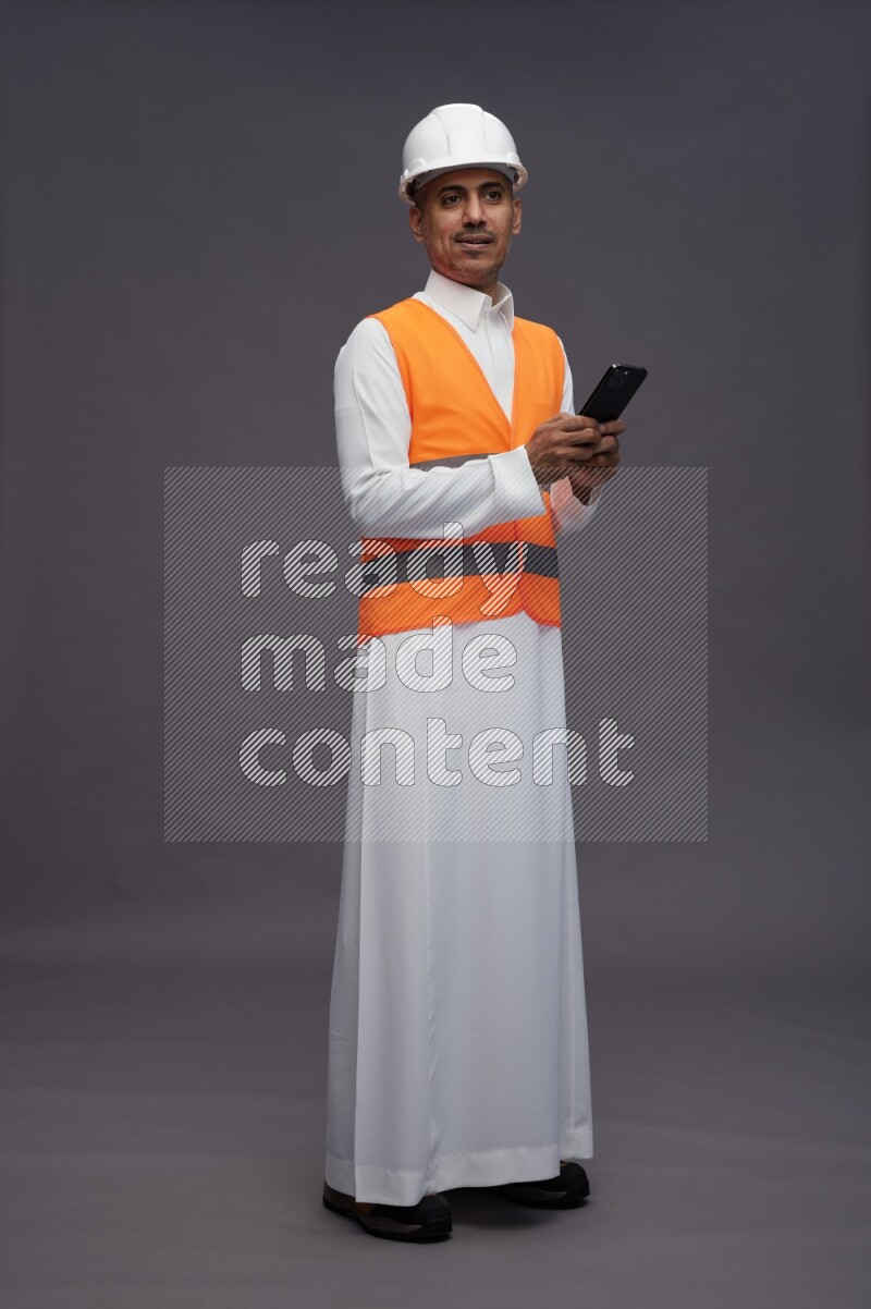 Saudi man wearing thob with engineer vest standing texting on phone on gray background