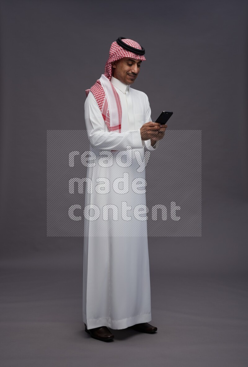 Saudi man wearing thob and shomag standing texting on phone on gray background