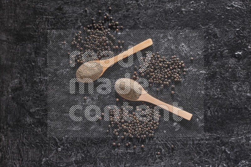 2 wooden spoons full of black pepper powder and black pepper beads spread on a textured black flooring