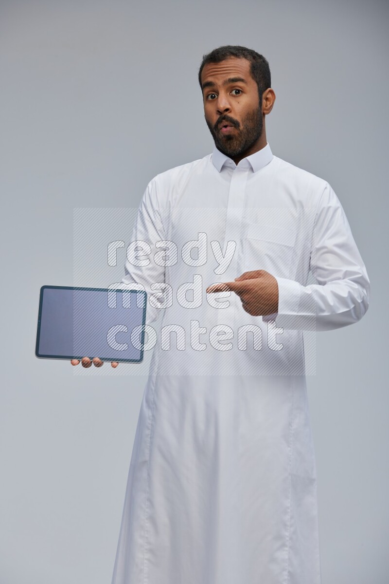 Saudi man wearing Thob standing showing tablet to camera on Gray background