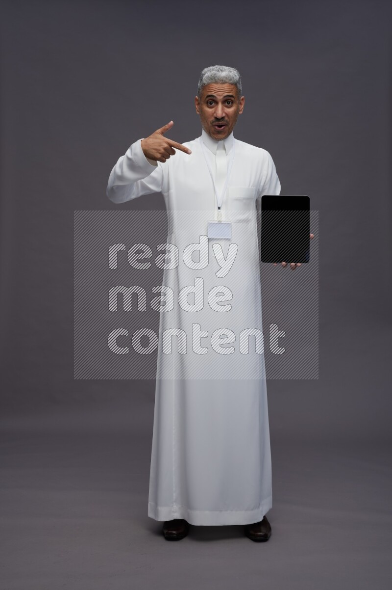 Saudi man wearing thob with neck strap employee badge standing showing tablet to camera on gray background