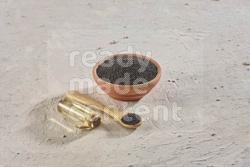 A wooden bowl and spoon full of black seeds with a bottle of black seeds oil on a textured white flooring