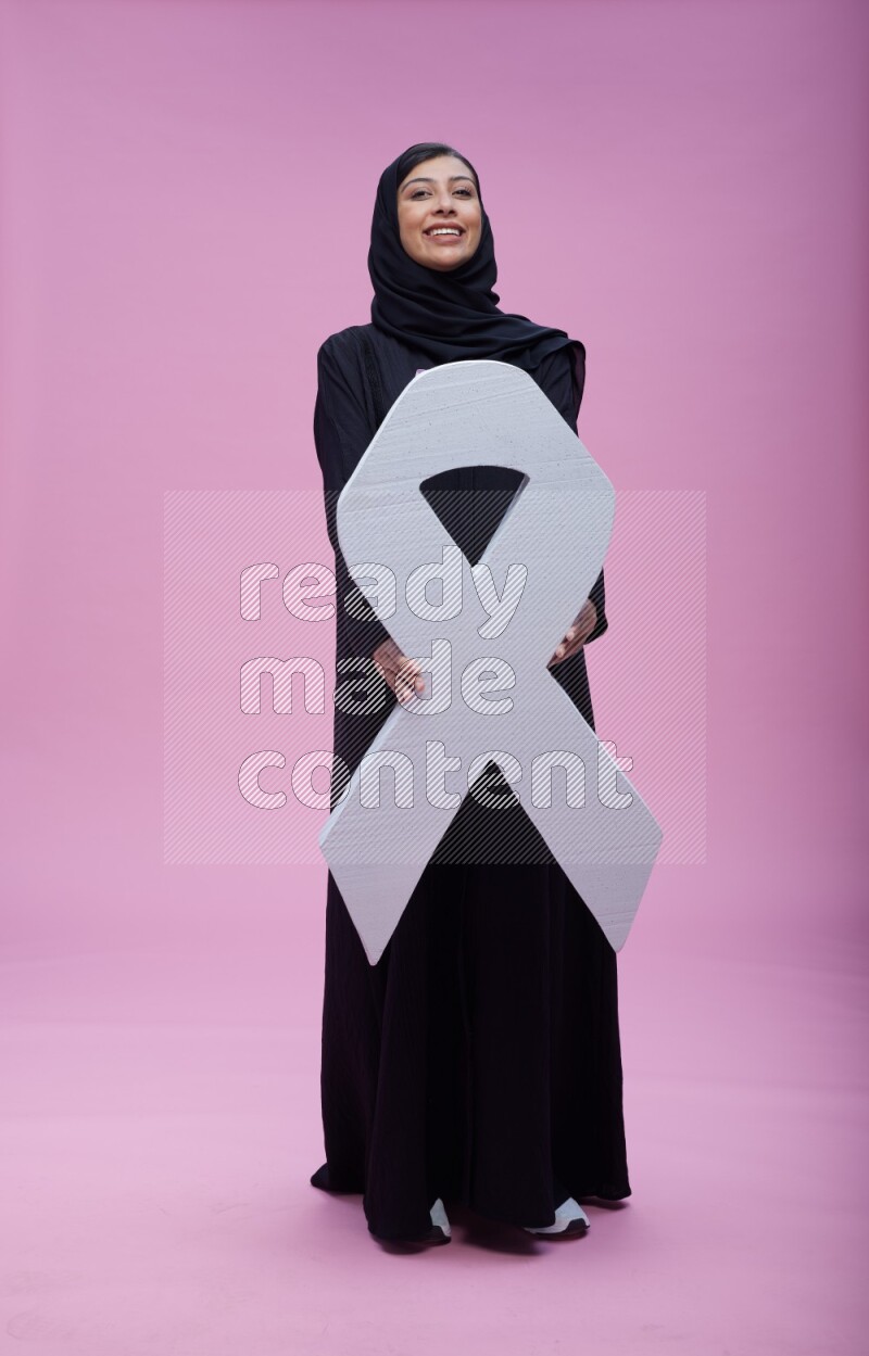Saudi woman wearing pink ribbon on abaya standing holding awareness ribbon on plain pink background