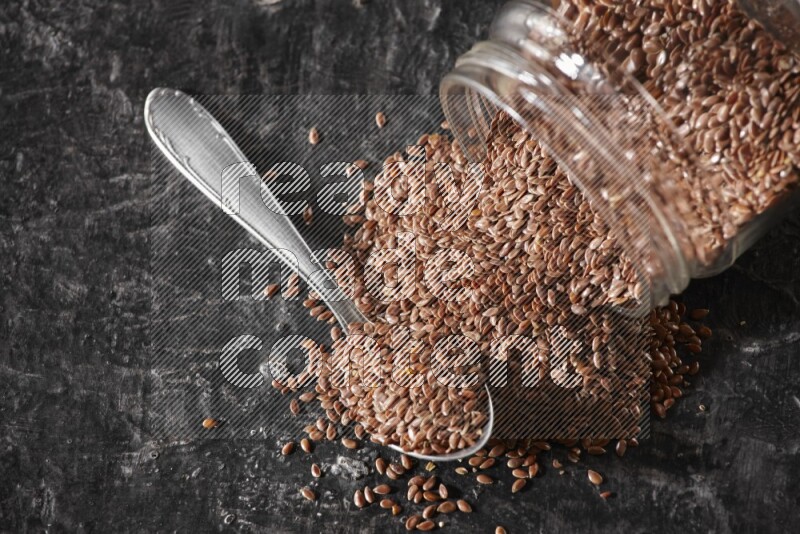 A glass jar full of flaxseeds flipped and seeds spread out with a metal spoon full of the seeds on a textured black flooring