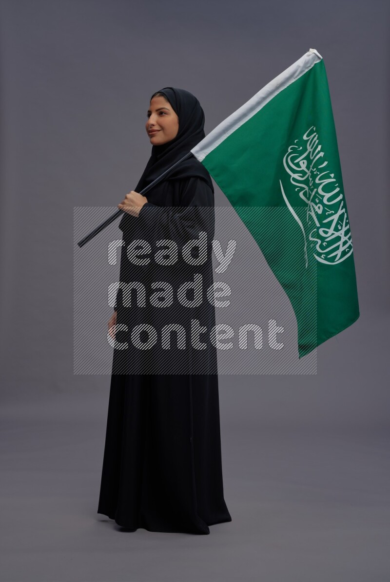 Saudi woman wearing Abaya standing holding Saudi flag on gray background