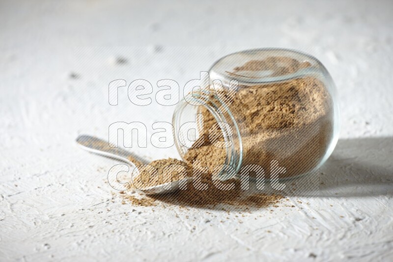 A flipped glass spice jar and a metal spoon full of cumin powder and powder spilled out on textured white flooring