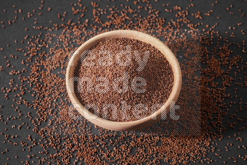 A wooden bowl full of garden cress seeds surrounded by seeds on a black flooring