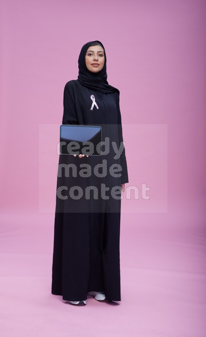 Saudi woman wearing pink ribbon on Abaya standing showing a tablet to camera on pink background