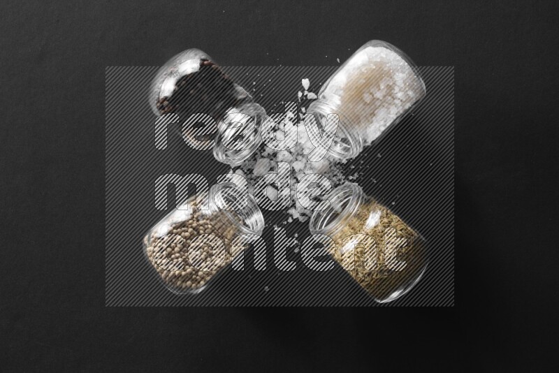 4 glass spice jars full of salt, black peppers, white peppers and cumin on black flooring